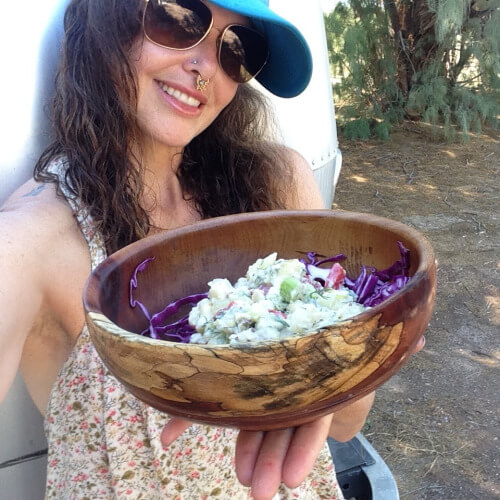 Melodia presenting a homemade festive potato salad in a handcrafted wooden bowl during an outdoor summer gathering.