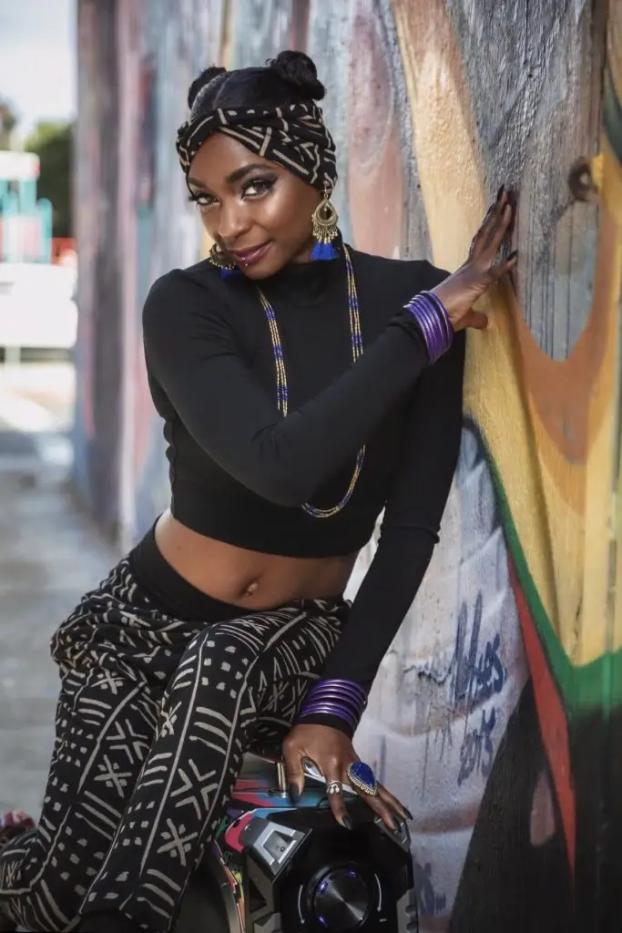 Model wearing black crop top and mudcloth print pants from the Mudcloth Collection, styled with headwrap and statement jewelry against urban mural backdrop.