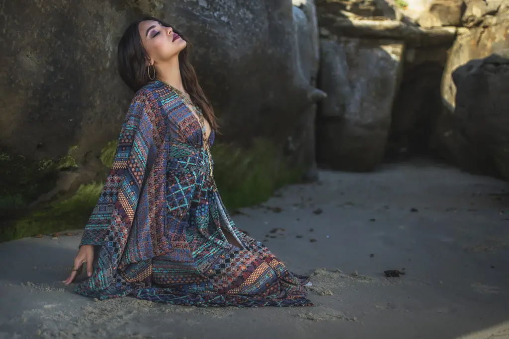 Our Mmdel Jade wearing a flowing bohemian maxi dress from the Bohemian Dreams Collection seated on sand between coastal rock formations.