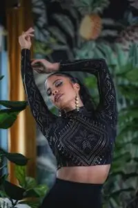 Model wearing a black long-sleeve crop top with gold geometric patterns, photographed indoors with lush greenery.