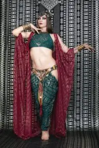 Model wearing a deep Malachite Assuit two-piece set with a fitted top and harem pants, styled with a flowing red sheer overlay and ornate belt.