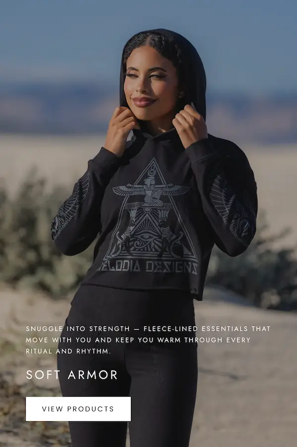 Model wearing a black Melodia Designs hoodie with a geometric emblem on the front, photographed outdoors in a desert setting.