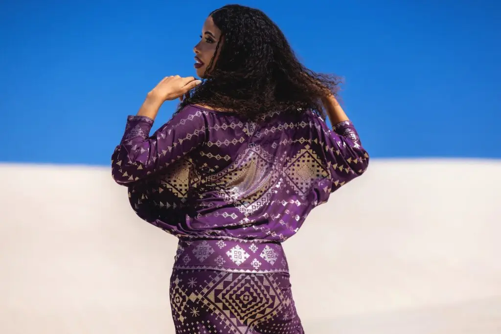 Model stands in white sand dunes wearing the Karnak Bella Batwing Blouse in aubergine — bold slow fashion for powerful movement.