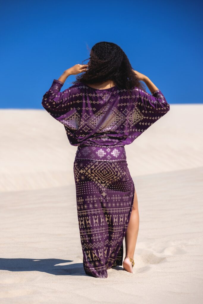 Back view of Karnak Bella Batwing blouse paired with the Alkimiya Cairo Split Skirt in aubergine, flowetry in motion across a sunlit dune landscape.