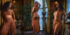 Model wearing a copper Karnak two-piece set with a fitted top, flowing harem pants, and a matching overlay dress, photographed indoors in a warm, ornate setting.