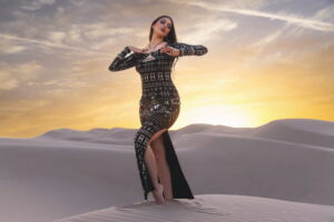 Woman wearing alkimiya nawal obsidian black sacred geometry dress posing on sand dunes at sunset