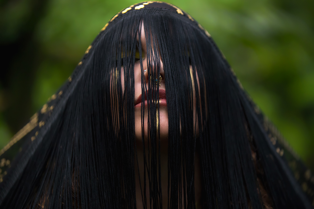 Portrait of Melodia with black fringe veil partially covering her face, featuring gold nose ring detail against a soft green background.