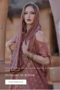 Model wearing a desert rose-toned Dendera Collection kaftan and matching hooded scarf with gold-inspired patterns, styled with stacked bangles against a warm architectural backdrop.